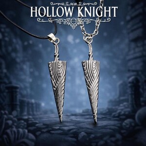 Puede incluir: Dos collares con colgantes plateados con diseño de daga. Los colgantes cuelgan de un cordón negro y una cadena plateada. El texto "HOLLOW KNIGHT" se muestra en la parte superior de la imagen.