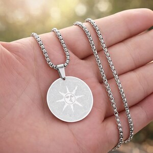 May include: Silver-toned necklace featuring a circular pendant with a sun design. The pendant has a detailed, etched pattern and is suspended from a matching chain. The necklace is shown against a blurred background.