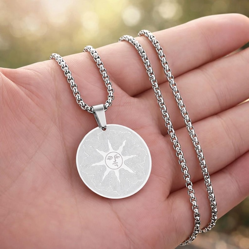 May include: Silver-toned necklace featuring a circular pendant with a sun design. The pendant has a detailed, etched pattern and is suspended from a matching chain. The necklace is shown against a blurred background.