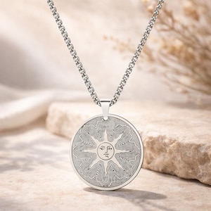 May include: A silver-toned pendant necklace featuring a circular pendant with a sun design. The sun has a face in the center, surrounded by radiating lines. The chain is a linked design. The necklace is displayed on a neutral-toned surface.