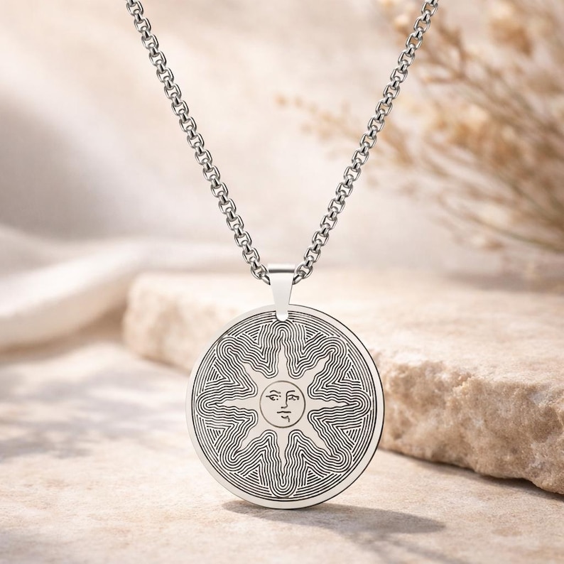 May include: A silver-toned pendant necklace featuring a circular pendant with a sun design. The sun has a face in the center, surrounded by radiating lines. The chain is a linked design. The necklace is displayed on a neutral-toned surface.