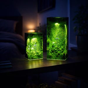 May include: Two illuminated resin art pieces. Each piece is a rectangular block with a green, moss-like interior. A silver sword is visible within the larger piece. The pieces are lit from within, creating a soft, green glow.