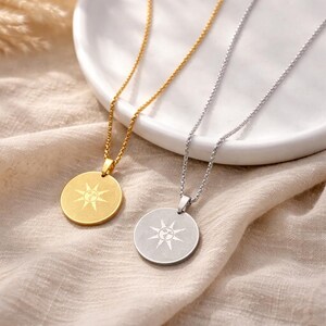 May include: Two necklaces with circular pendants, one gold-toned and one silver-toned, each featuring a sunburst design. The pendants hang from delicate chains. The necklaces rest on a beige fabric and a white plate.