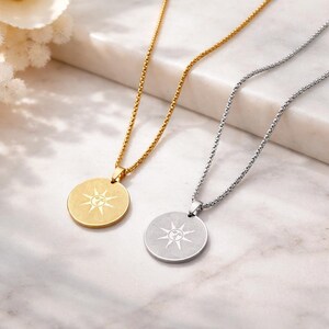 May include: Two necklaces with round pendants. One is gold-toned, the other silver-toned. Each pendant features a sunburst design. The chains are delicate and the pendants are centered. The background is a marble surface.