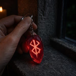 Puede incluir: Un llavero rojo translúcido con forma de cristal irregular que presenta el símbolo blanco brillante de la Marca del Sacrificio grabado en el centro. El colgante está unido a una anilla de metal plateado mediante una cadena corta.