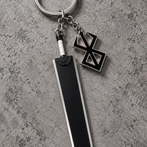 May include: A metal keychain featuring a large, flat, black sword pendant with a white handle wrap and a separate geometric black and silver charm shaped like a stylized brand symbol, all attached to a circular silver split ring.