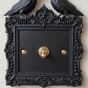 May include: Black light switch cover with two black crow figurines above the frame. The frame has an ornate, scrollwork design. The switch and screws are gold-coloured.
