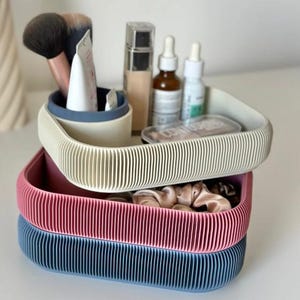May include: Three ribbed storage trays in beige, pink, and blue, stacked on top of each other. The top tray holds makeup brushes and bottles. The middle tray contains scrunchies. The bottom tray is empty. The trays are on a white surface.