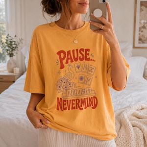 May include: A mustard yellow short-sleeve t-shirt featuring a graphic print with the text "PAUSE... NEVERMIND" in red. The design includes a blue peacock illustration and several mahjong tiles arranged in a cluster and on a rack.