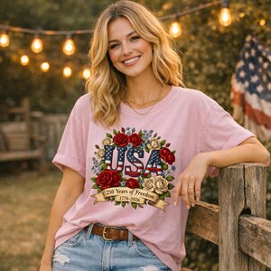 Op de afbeelding: Lichtroze t-shirt met een bloemenontwerp met het woord "USA" in rood, wit en blauw. Het ontwerp bevat rode en witte rozen en een banner met de tekst "250 Years of Freedom 1776-2026".