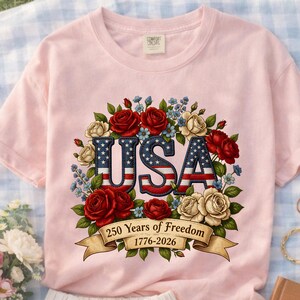 Op de afbeelding: Lichtroze T-shirt met het woord "USA" in een patriottisch ontwerp. De letters zijn omgeven door rode en witte rozen, blauwe bloemen en een banner met de tekst "250 Years of Freedom 1776-2026".