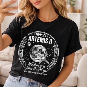 May include: Black t-shirt featuring a NASA Artemis II design. The graphic includes an illustration of the moon, a spacecraft, and the text "We love you from the Moon." The shirt also includes the date "APRIL 2026 - AROUND THE MOON & BACK."