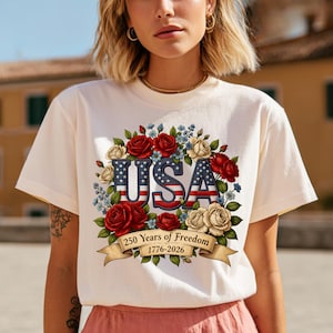 Op de afbeelding: Cr&egrave;mekleurig T-shirt met een bloemenontwerp met het woord "USA" in rood, wit en blauw. Het ontwerp bevat rode en cr&egrave;merozen, blauwe bloemen en een banner met "250 Years of Freedom 1776-2026".