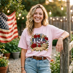 Op de afbeelding: Lichtroze t-shirt met een bloemenontwerp en het woord "USA" in rood, wit en blauw. Het ontwerp bevat rode rozen en de tekst "250 Years of Freedom 1776-2026". Het shirt wordt gedragen met een spijkerbroek en een bruine riem.