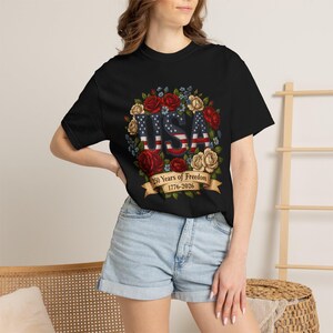 Op de afbeelding: Zwart T-shirt met een bloemenkransontwerp en het woord "USA" in een patriottisch lettertype. De letters zijn gevuld met een sterren- en strepenpatroon. Een banner hieronder luidt "250 Years of Freedom 1776-2026".