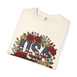 Op de afbeelding: Cr&egrave;mekleurig T-shirt met het woord "USA" in een patriottisch ontwerp. De letters zijn gevuld met een denim-achtige textuur en de Amerikaanse vlag. Het woord is omgeven door rode, cr&egrave;me en blauwe bloemen.