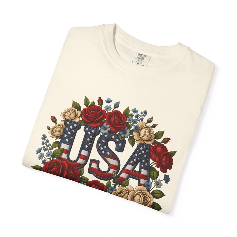 Op de afbeelding: Cr&egrave;mekleurig T-shirt met het woord "USA" in een patriottisch ontwerp. De letters zijn gevuld met een denim-achtige textuur en de Amerikaanse vlag. Het woord is omgeven door rode, cr&egrave;me en blauwe bloemen.