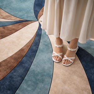 May include: A close-up of a rug with a spiral design in shades of blue, brown, and beige. White sandals with braided straps are visible. The rug's pattern creates a sense of movement and visual interest.