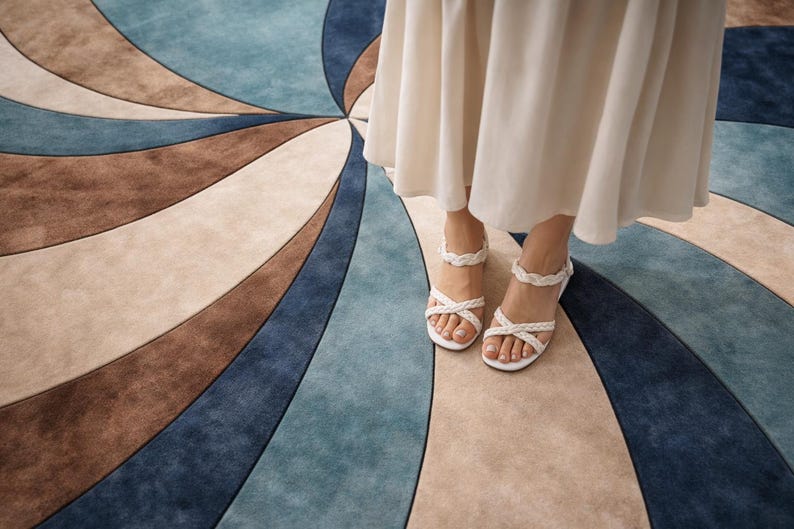 May include: A close-up of a rug with a spiral design in shades of blue, brown, and beige. White sandals with braided straps are visible. The rug's pattern creates a sense of movement and visual interest.