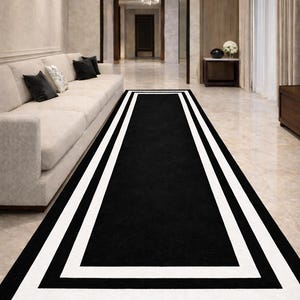 Peut inclure: Un tapis rectangulaire noir avec une bordure blanche et deux bandes blanches. Le tapis est placé dans un couloir avec un canapé crème, des coussins noirs et un lustre. Les dimensions du tapis conviennent à un long couloir.