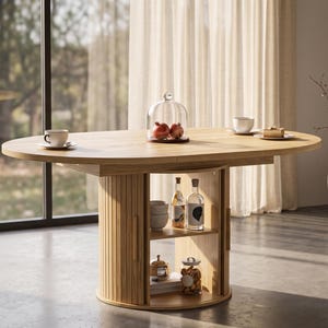 May include: A light oak oval dining table with a central pedestal base featuring vertical slats and open shelving. The tabletop holds a glass cloche with pomegranates, a white teacup, and a small dessert plate. The shelves contain glass bottles and jars.