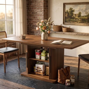 May include: A rectangular wooden dining table with a central fluted pedestal base featuring open storage shelves, accompanied by two chairs with cane backrests and black seats. The table surface holds a ceramic vase with flowers and an open book.