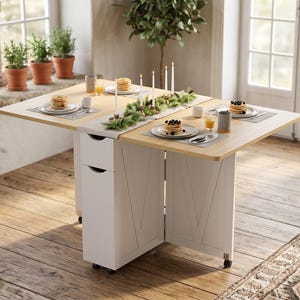 May include: A light wood-toned drop-leaf dining table with a white base featuring two integrated storage drawers. The table is set with plates of pancakes, glasses of juice, mugs, and a central table runner decorated with pine branches and candles.