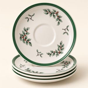 Puede incluir: Una pila de cuatro platillos de cerámica blanca con un borde verde oscuro y un patrón repetitivo de hojas de acebo verdes con pequeñas bayas rojas. Cada platillo tiene una hendidura circular central para una taza.