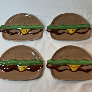 May include: Four ceramic plates shaped like hamburgers. Each plate features a brown bun, green lettuce, brown patty, and yellow cheese. The plates are arranged on a white surface.