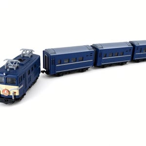 May include: A blue model electric locomotive train engine with a cream-coloured front panel and a circular red emblem, followed by three matching blue passenger train cars. Each car features rectangular windows and a grey roof, connected in a line.