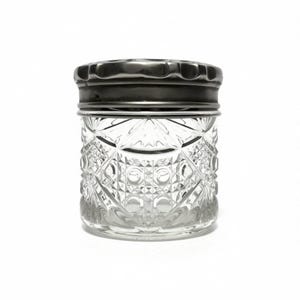 Op de afbeelding: Een doorzichtige glazen pot met een cilindrisch lichaam voorzien van een gedetailleerd ruit- en kruisarceringpatroon. De pot is afgesloten met een donker, metalen, rond deksel met een geschulpte rand en een gladde, licht reflecterende afwerking.