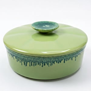 May include: A round, light green ceramic casserole dish with a matching lid. The lid features a small, circular handle with a dark green, mottled glaze. The upper rim of the base has a decorative, dark green drip glaze effect over the light green.
