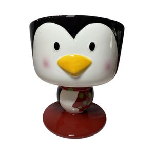 Può includere: Un contenitore a forma di pinguino in ceramica con parte superiore nera, viso bianco, becco giallo e base rossa. Il pinguino ha occhi neri e guance rosee. Il contenitore è un design stravagante, perfetto per piccoli oggetti o come decorazione.
