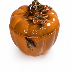 Puede incluir: Un tarro de cerámica naranja brillante con forma de calabaza y tapa, que presenta un tallo esculpido y hojas detalladas en tonos marrón y verde. La superficie tiene un acabado liso y reflectante con surcos verticales definidos.