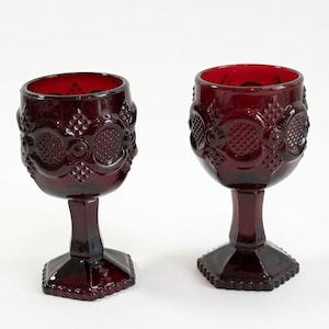 May include: Two deep red glass goblets featuring a textured pattern of raised circles and diamond shapes on the bowl. Each goblet has a thick, faceted stem and a hexagonal base with a beaded edge, crafted from translucent ruby-colored glass.