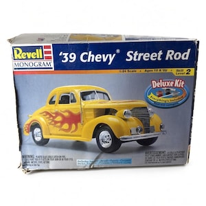 May include: A Revell Monogram '39 Chevy Street Rod model kit box. The box features a yellow car with red flame decals, a 1:24 scale, skill level 2, and a Deluxe Kit label indicating that paint, glue, and a brush are included in the package.