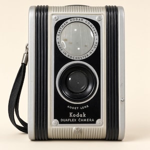 Op de afbeelding: Een vintage Kodak Duaflex camera met een zilveren en zwarte behuizing, voorzien van een Kodet-lens en een zwarte polsband. De voorplaat toont de tekst Eastman Kodak Company Made in Rochester NY USA en Kodak Duaflex Camera in het wit.