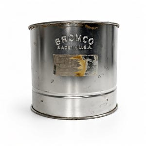 May include: A cylindrical metal flour sifter with a silver finish. The exterior features the embossed text BROMCO MADE IN U.S.A. and a rectangular label with a partially torn, aged paper surface showing remnants of dark ink and adhesive residue.