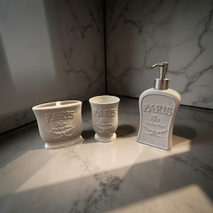 Puede incluir: Un juego de baño de tres piezas en color gris claro. El juego incluye un porta cepillos de dientes, un vaso y un dispensador de jabón. Cada pieza está grabada con la palabra "PARIS" y elementos florales decorativos.