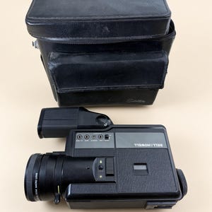 May include: A black Bell & Howell movie camera with a zoom macro lens and a side-mounted handle, accompanied by a matching black rectangular carrying case with the word Satellite embossed on the front flap. The camera features various control ports.