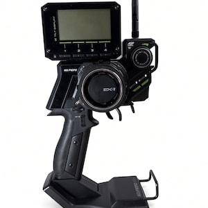 Puede incluir: Un transmisor de radiocontrol negro KO Propo EX-1 con una pantalla inclinable montada en la parte superior, un volante, una antena vertical y un mango tipo pistola. El dispositivo está montado sobre una base de plástico negro con la marca.