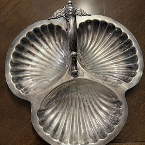 May include: A silver-colored, three-section serving dish with a handle. The dish has a shell-like design in each section and a decorative handle. The dish is made of metal and is likely vintage.