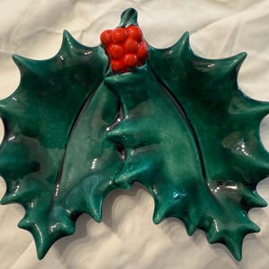 May include: A ceramic holly leaf-shaped dish with a glossy green finish. The dish features a cluster of red berries at the top. The edges of the leaves are pointed. This decorative piece is suitable for holiday decor.