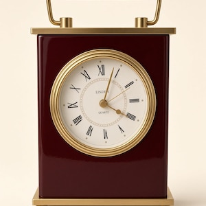May include: A rectangular table clock with a deep burgundy finish and a gold-toned metal frame. The clock face is off-white with black Roman numerals, gold hands, and the text LINDEN QUARTZ. It features a gold handle on top and four small feet.