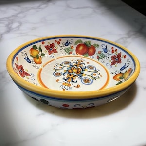 May include: A decorative ceramic bowl with a yellow rim and a hand-painted floral and fruit design. The bowl features a central floral motif and is adorned with red, orange, and blue accents. The bowl is round and sits on a white surface.