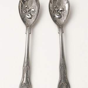 May include: A silver-toned metal salad serving set consisting of a fork and a spoon. Both utensils feature an embossed design of fruit and leaves in the bowl and a decorative shell-like motif at the base of the handle with vertical lines.