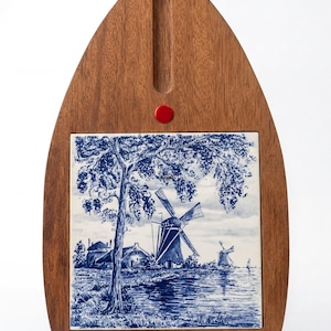 Può includere: Un tagliere in legno a punta con una piastrella quadrata in ceramica centrale raffigurante un'illustrazione blu e bianca di un paesaggio olandese, inclusi mulini a vento, un albero e uno specchio d'acqua. Un piccolo accento circolare rosso.