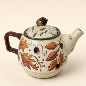 May include: A cream-coloured ceramic teapot featuring a painted autumn motif of orange and brown oak leaves, acorns, and small red berries. The lid is topped with a sculpted acorn handle, and the teapot has a dark brown handle and a spout.