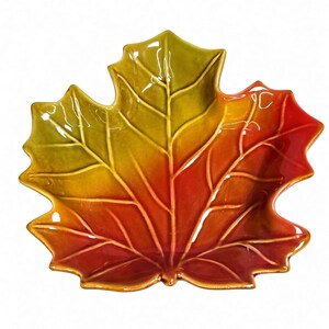 May include: A ceramic dish shaped like a maple leaf with a glossy finish. The surface features a gradient colour transition from a vibrant yellow-green at the top to a deep, warm orange and red at the base, with raised veins defining the leaf structure.