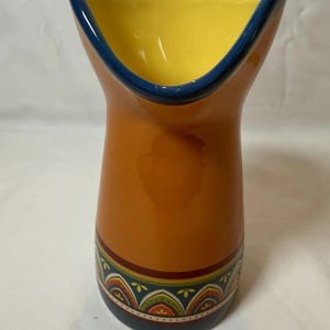 May include: A ceramic pitcher with a wide, curved opening. The interior is yellow, the rim is outlined in blue, and the exterior is orange. A decorative band with a floral pattern encircles the base. The pitcher is approximately 8 inches tall.